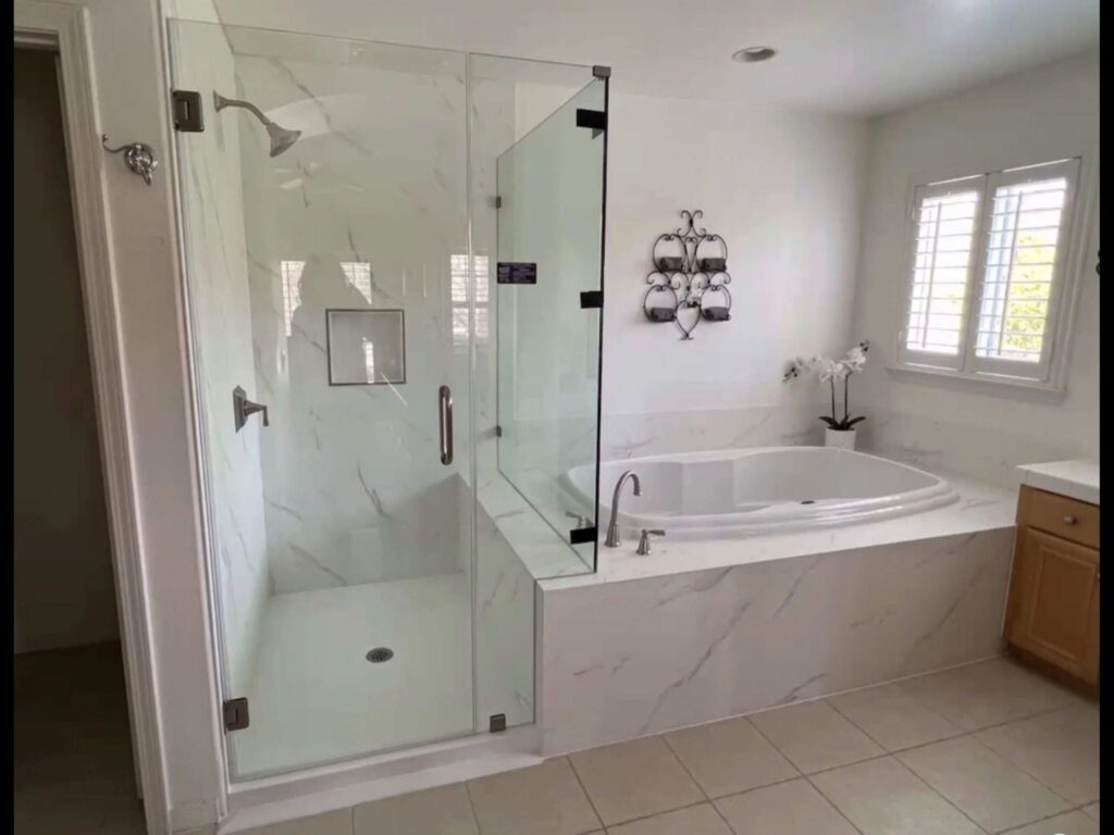 Professionally remodeled shower in Philadelphia featuring a frameless glass shower enclosure with white marble tile walls, built-in niche, corner soaking tub with marble surround, chrome fixtures, and natural light from shuttered windows.